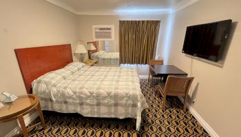 A hotel room with a bed, TV, desk, chairs, and lamp. The decor includes patterned carpeting and curtains, and a wall-mounted air conditioner.