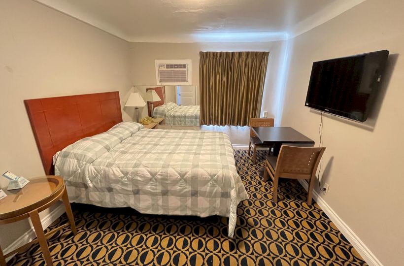 A hotel room with a bed, TV, desk, chairs, and lamp. The decor includes patterned carpeting and curtains, and a wall-mounted air conditioner.