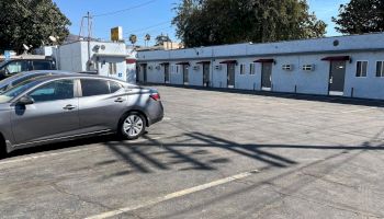 A small parking lot with a few cars is in front of a row of single-story motel rooms. The sky is clear, and trees are visible in the background.