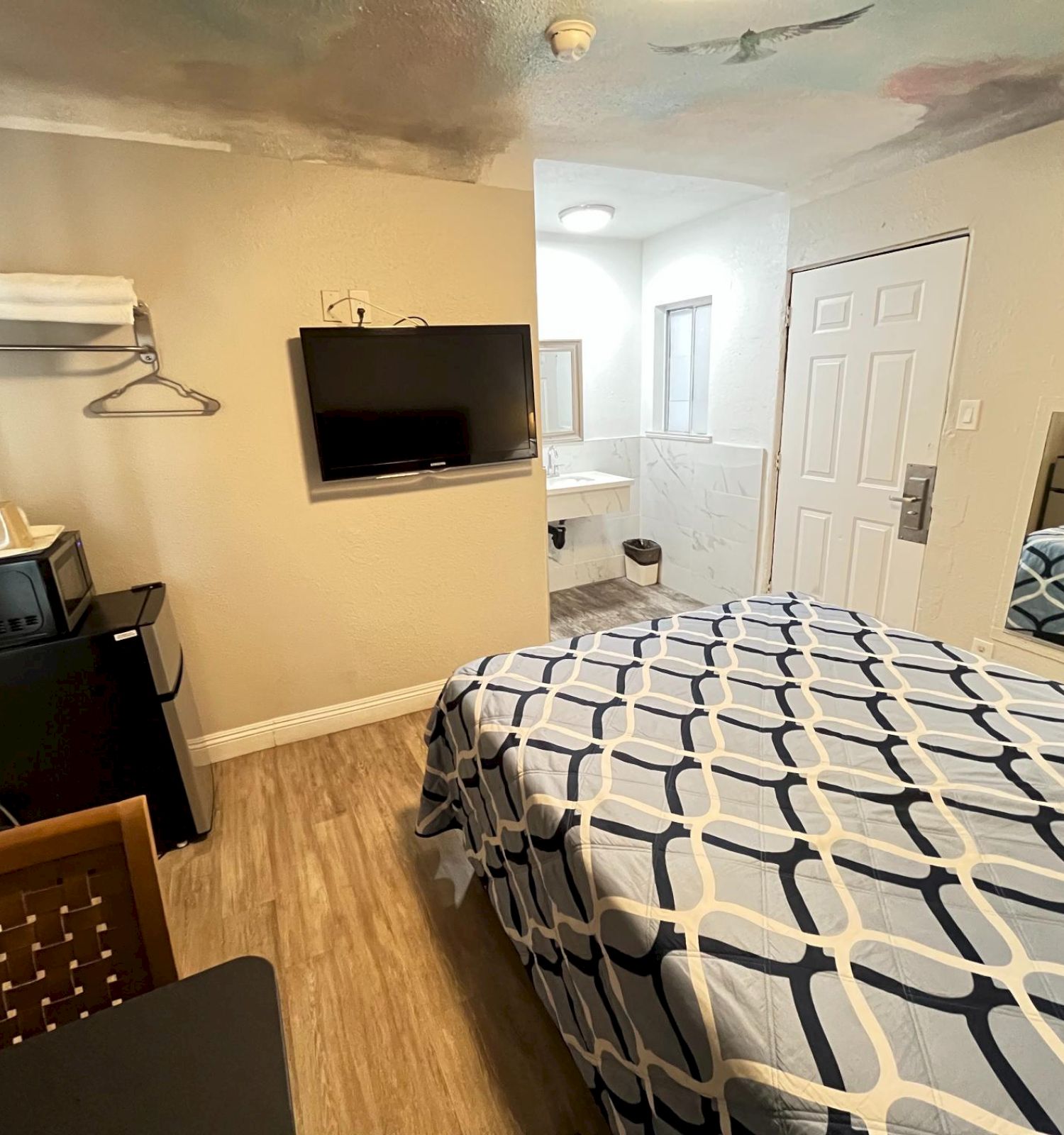 A small room with a patterned bed, wall-mounted TV, mini fridge, open closet, and bathroom. Light wood floor and neutral walls complete the setup.