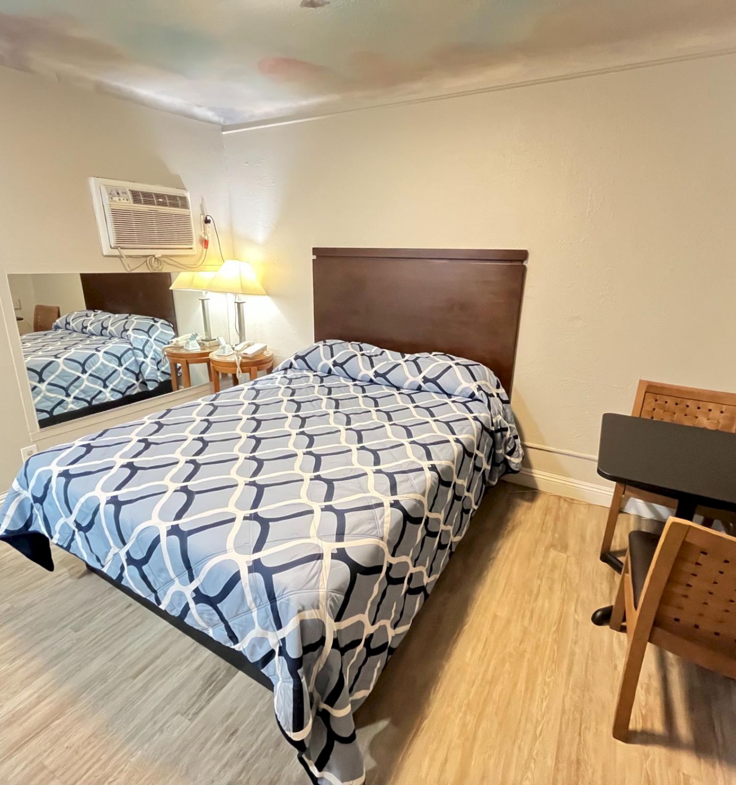 A small hotel room with a bed, table, chairs, lamp, air conditioning, and mirror on the wall.