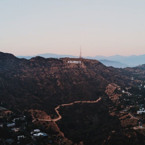 The image shows an aerial view of the Hollywood Hills with the iconic Hollywood sign visible on the hillside and scattered homes below.