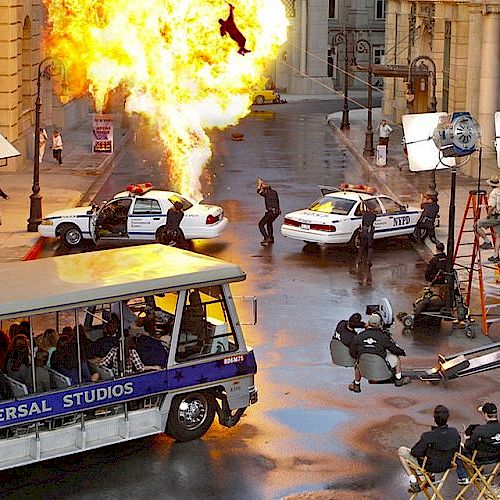 The image shows a film set with an explosion, police cars, crew, and a Universal Studios tour bus observing the action.